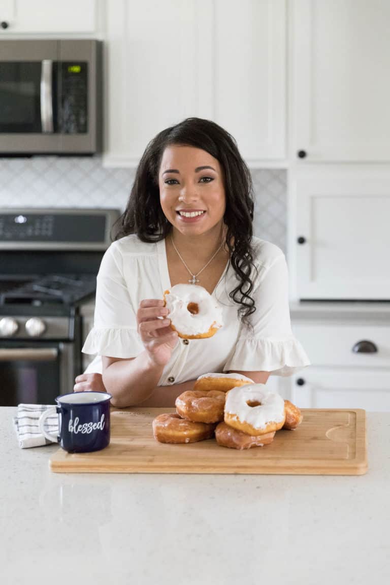 Rich, fluffy glazed donuts with white icing and a caramel center on a wooden cutting board in a modern white kitchen. A smiling woman holds a donut, showcasing the irresistible treat from sweetsherrypie.com.