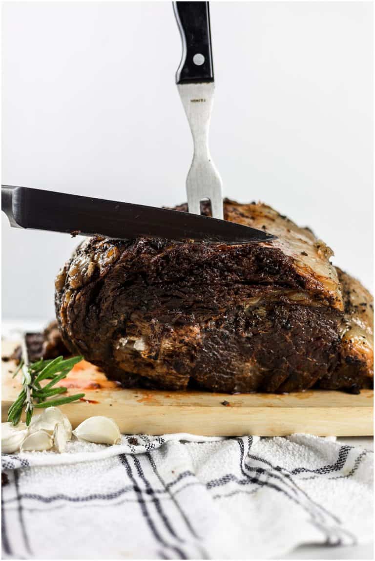 Juicy roasted beef being carved on a wooden cutting board with a carving knife and fork, garnished with garlic and fresh rosemary on a white and black checkered cloth, perfect for a hearty meal.