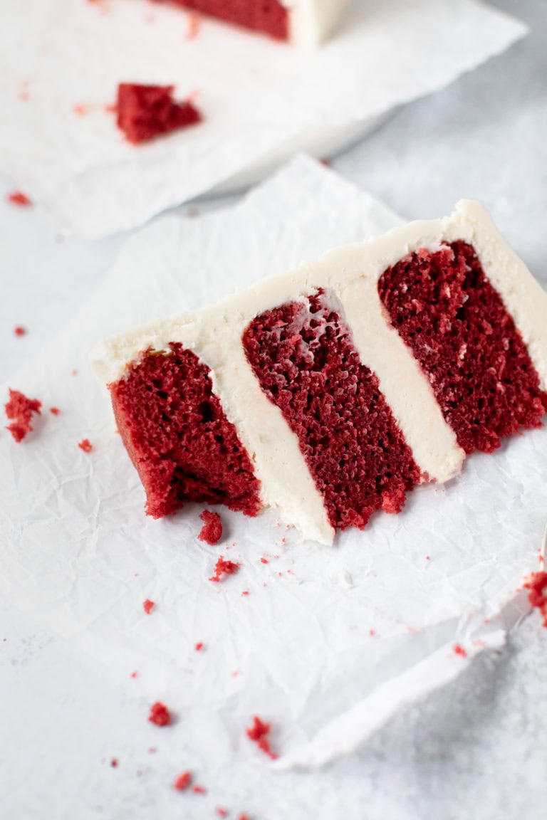 Rich red velvet cake with cream cheese frosting, sliced and presented on white parchment paper, showcasing the moist texture and vibrant color. Perfect for special occasions or classic desserts.
