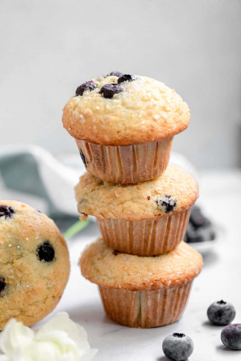 Fresh blueberry muffins stacked on a white marble surface with blueberries scattered around, showcasing delicious homemade blueberry muffins with moist texture and ripe blueberries, perfect for breakfast or snacks.