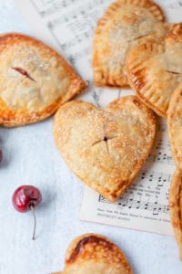 Sweetheart-shaped breakfast pastries with flaky crust and a sprinkle of sugar, served on parchment paper with a cherry and hymn sheet for a cozy, homemade breakfast vibe.
