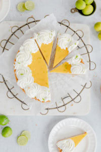 Lemon pie with whipped cream and lime zest on top, served on a wire cooling rack. Fresh lime slices and whole limes surround the dessert, highlighting citrus flavors. Perfect for summer gatherings or desserts.