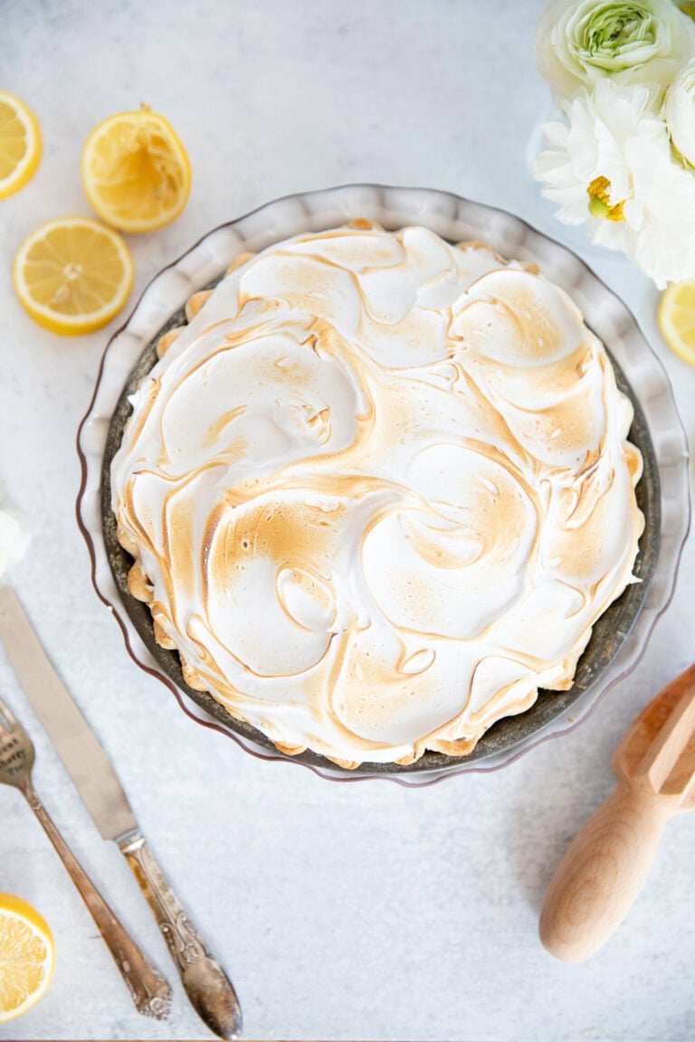 Lemon meringue pie topped with toasted meringue, garnished with fresh lemon slices and flowers, served on a white surface, perfect for lemon dessert lovers.