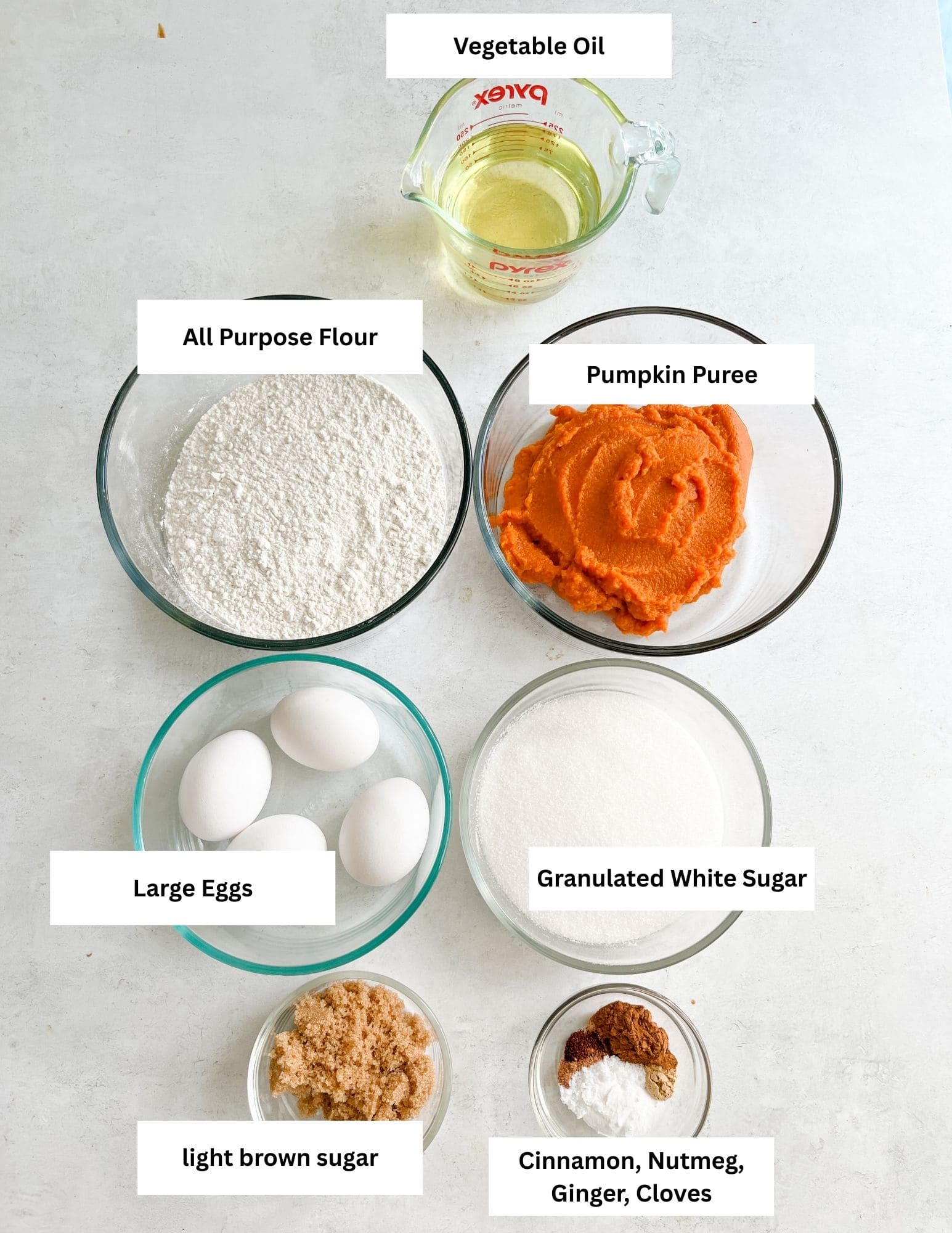 Bright ingredients for pumpkin pie baking including large eggs, granulated white sugar, all purpose flour, pumpkin puree, vegetable oil, light brown sugar, spices like cinnamon, nutmeg, ginger, cloves, and measuring cups on a white surface.