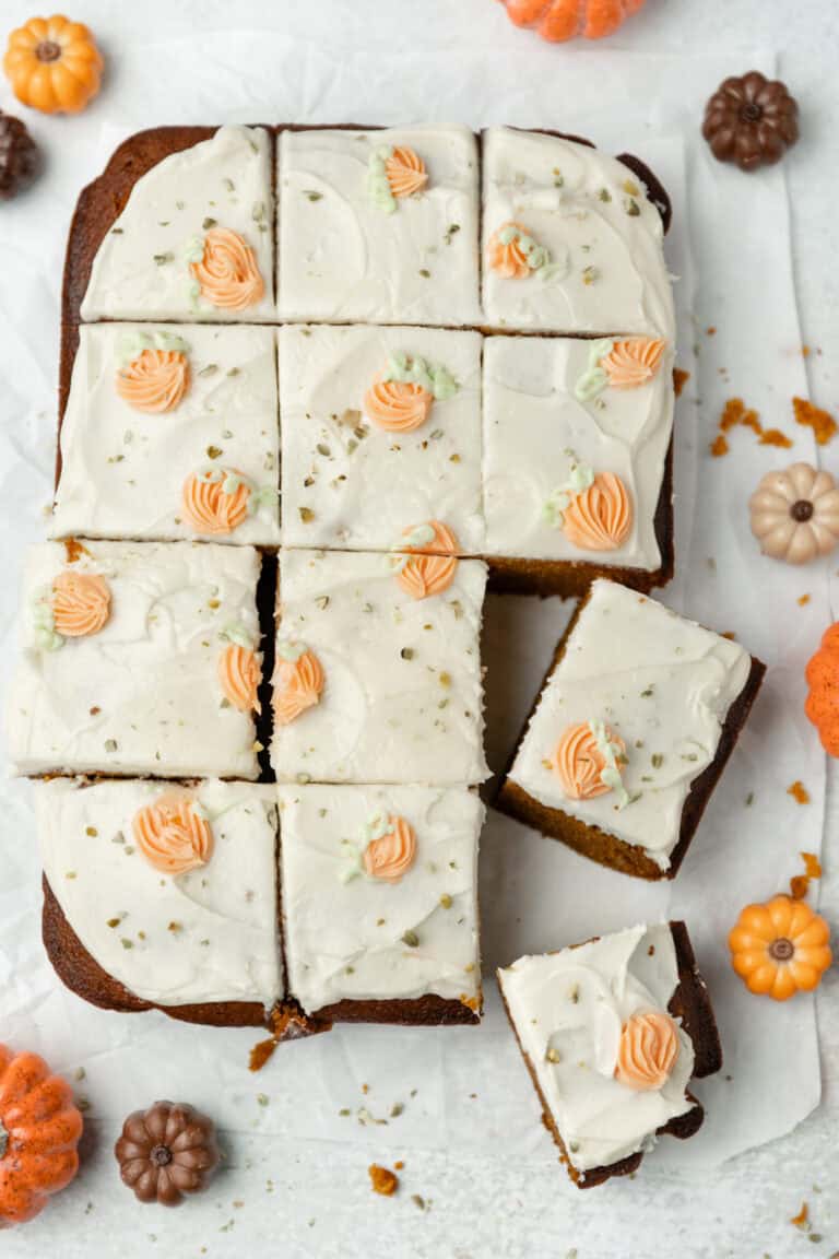 Delicious pumpkin cake with cream cheese frosting topped with orange and green piped icing flowers, surrounded by mini pumpkins for fall dessert.